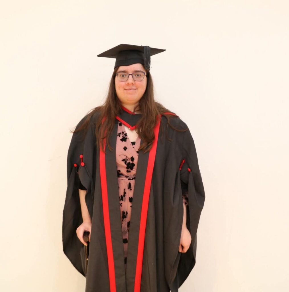 A photo of Laura Ann Moulding in a red and black graduation gown, with academic cap. Underneath the gown I am wearing a pink and black dress.